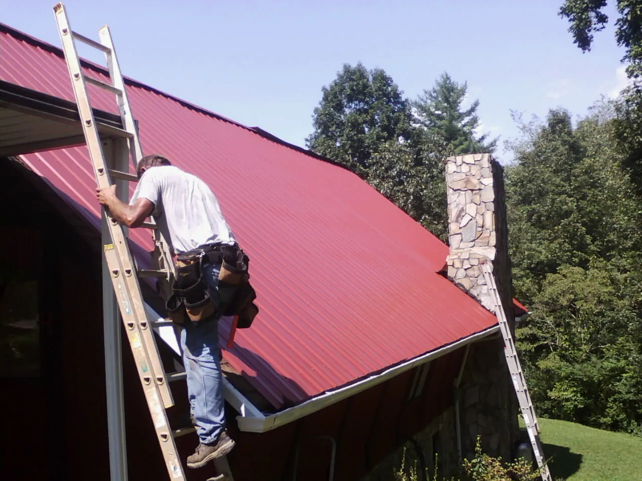 Roofer on a ladder beside a red metal roof and stone chimney — Soffit & Fascia Repair by Roofing Brothers in Warm Springs